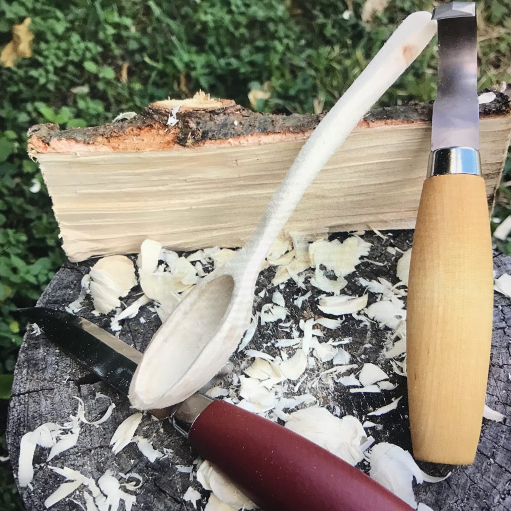 A short split log, two carving knives and a wood spoon sitting on a tree stump