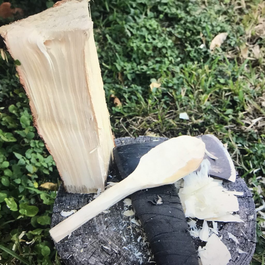 A short piece of slit pine and a rough shaped wood spoon resting on an hand axe all sitting on a tree stump