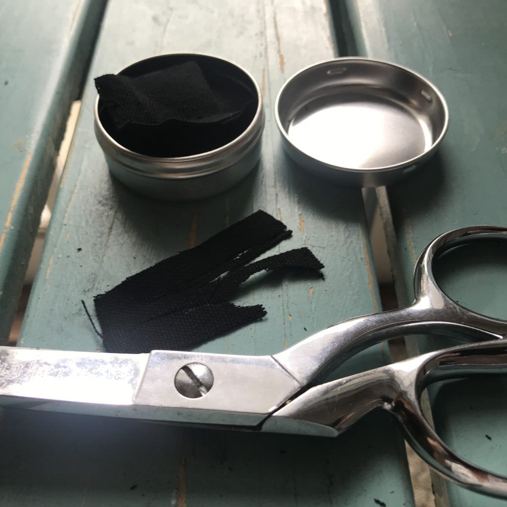A small metal tin with char cloth inside. A small price of char cloth and a pair of scissors on a table