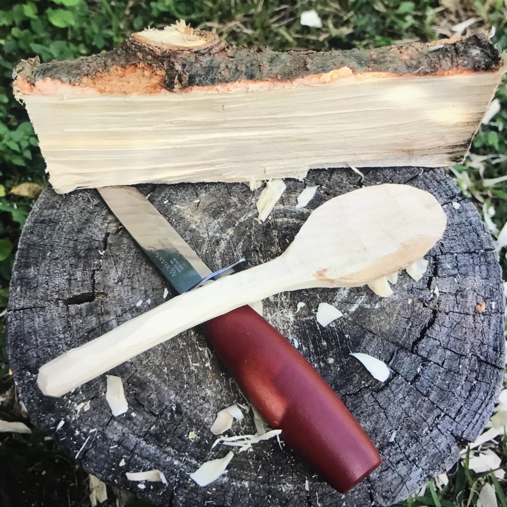 A roughly shaped wood spoon laying on a tree stump with a wood carving knife sitting on a tree stump