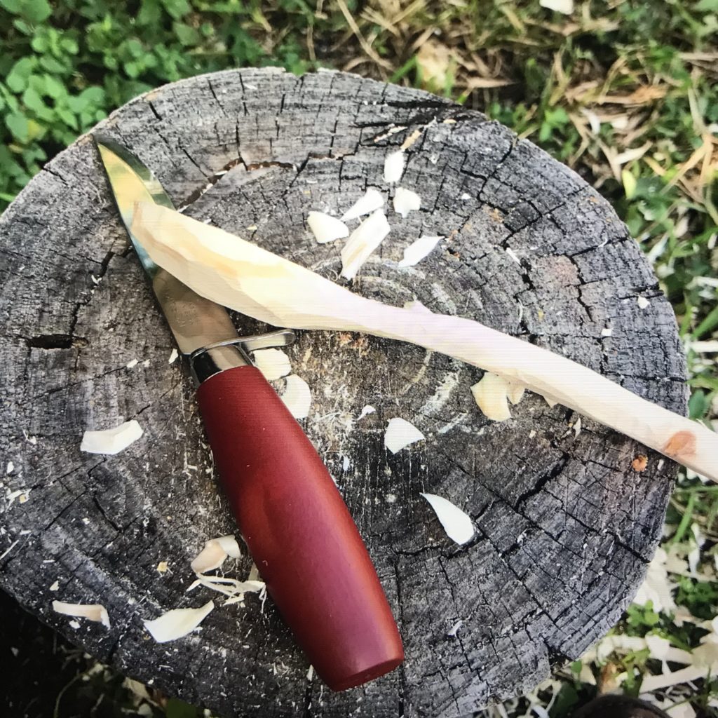 Side profile of a wood spoon along side a wood carving knives sitting on a stump