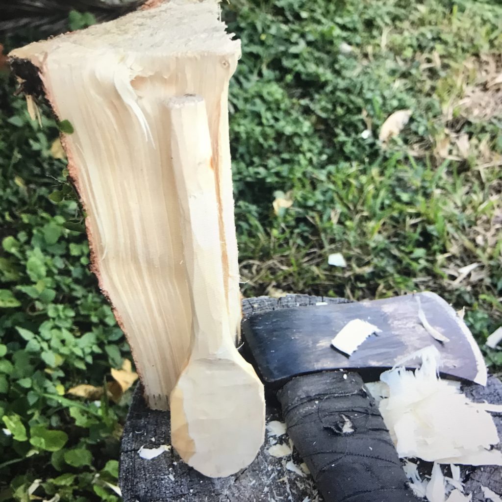 A rough shaped wood spoon leaning up against a short piece of split log and a hand axe sitting on a tree stump