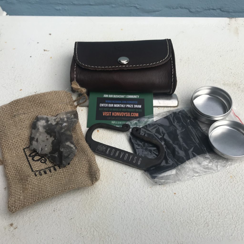 A small leather pouch with it contents displayed including a metal sticker a small tin a flint stone and some char cloth with small burlap sack