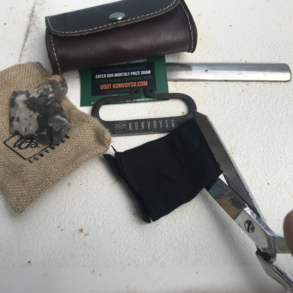 A small leather pouch with it contents displayed including a metal sticker a small tin a flint stone and some char cloth with small burlap sack With scissors cutting char cloth